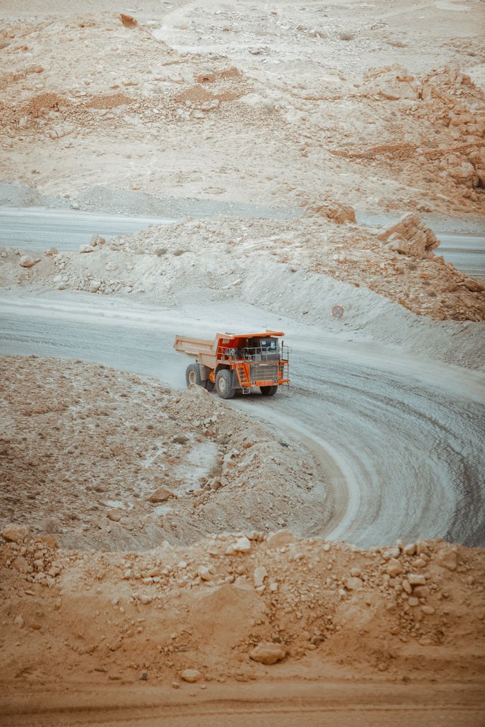 A heavy dump truck navigates a winding dirt road through the barren desert landscape of Biskra, Algeria.